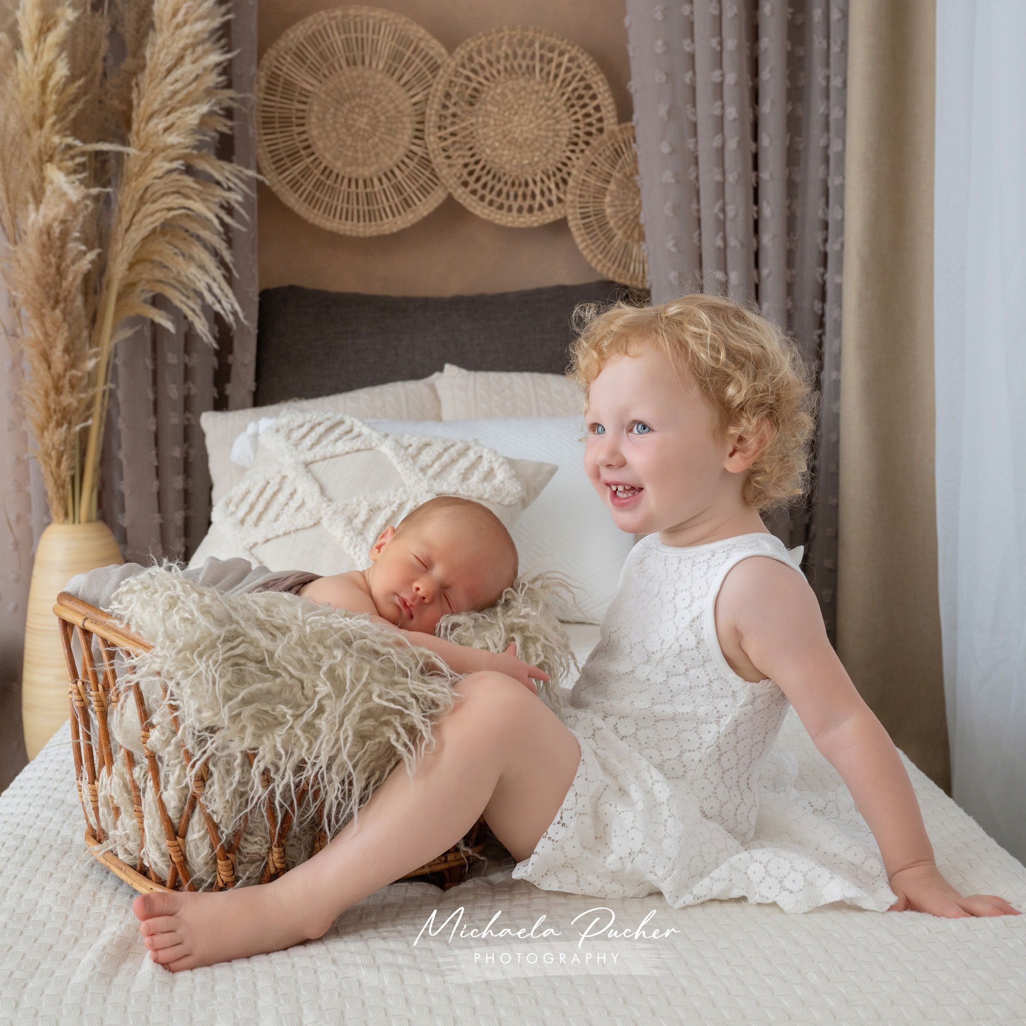 Älteres Geschwisterkind sitzt lachend auf dem Bett, während das Neugeborene friedlich in einem kleinen Korb daneben schläft; umgeben von warmen Boho-Deko-Elementen.